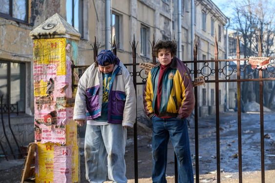 An screenshot of the movie Grom: Trudnoe Destyovo, as two kids in 90s clothings are held up by their collars on a fence